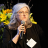 Photo of Thora Karadottir speaking onstage with a bouquet behind her