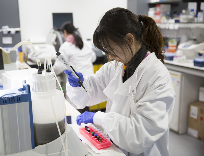 A young woman CSCI researcher pipetting in the lab, with another researcher in the background at the lab bench